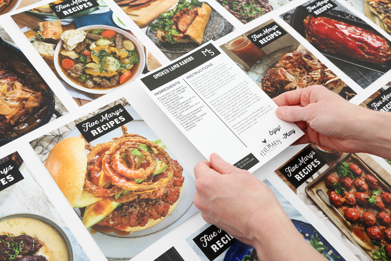 Two hands holding a recipe card with more recipe cards surrounding it.