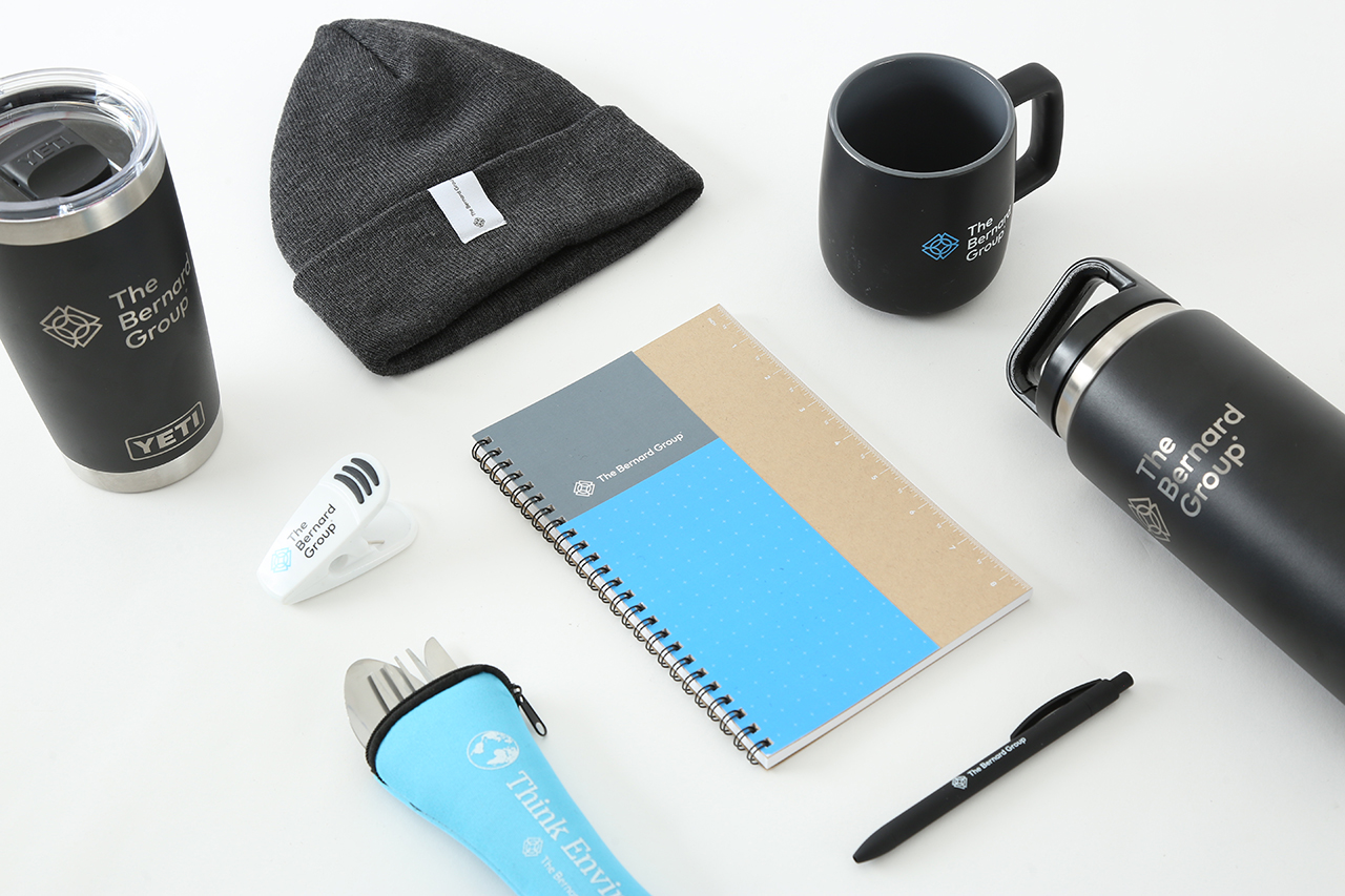 A collection of The Bernard Group branded items, including a notepad, mugs, hat, chip clip and silverware.