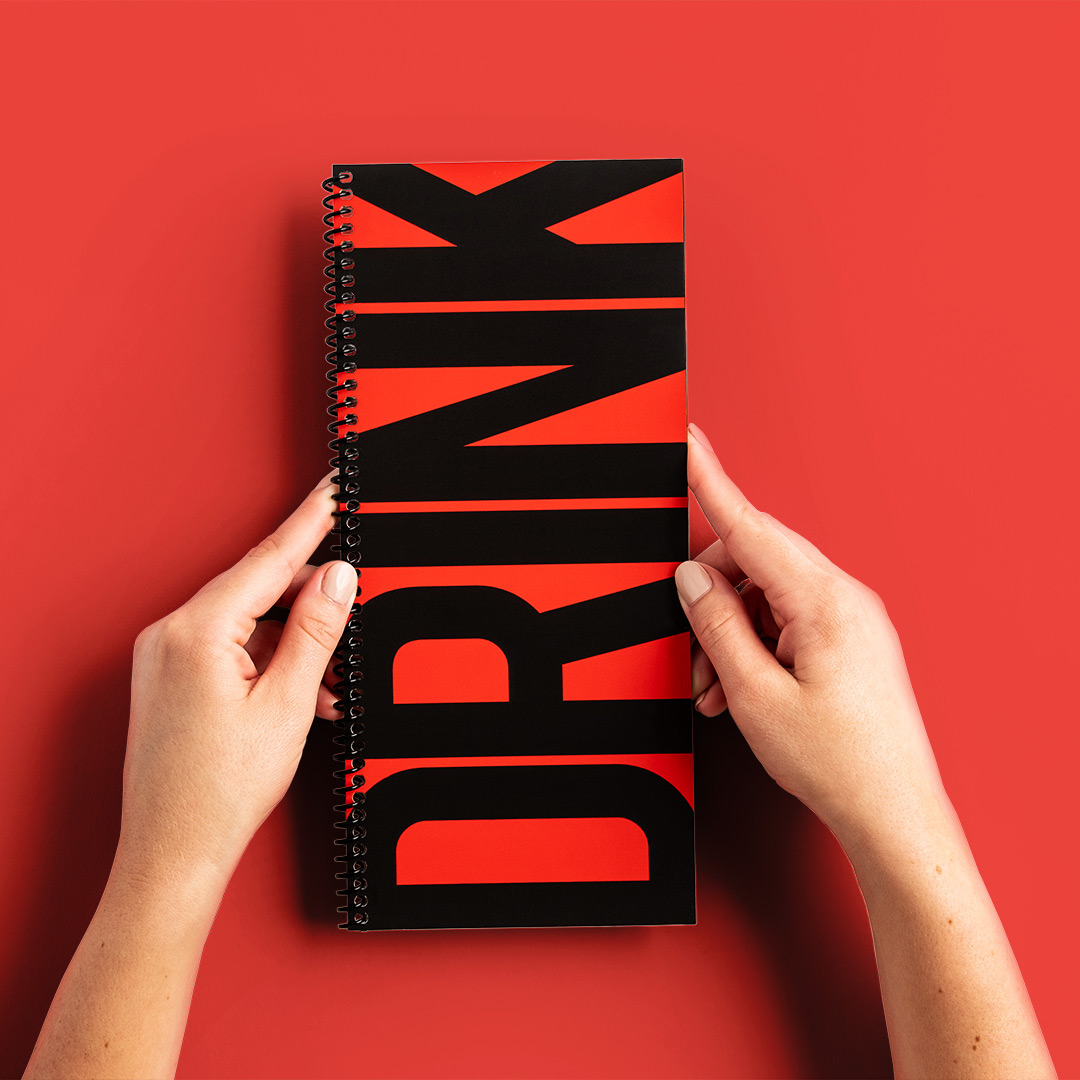 Two hands holding a drink menu with a red and black design and a spiral binding.