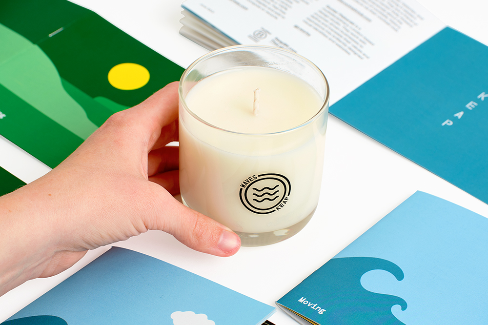 A hand holding a Keap Waves candle with custom product catalogs around it.