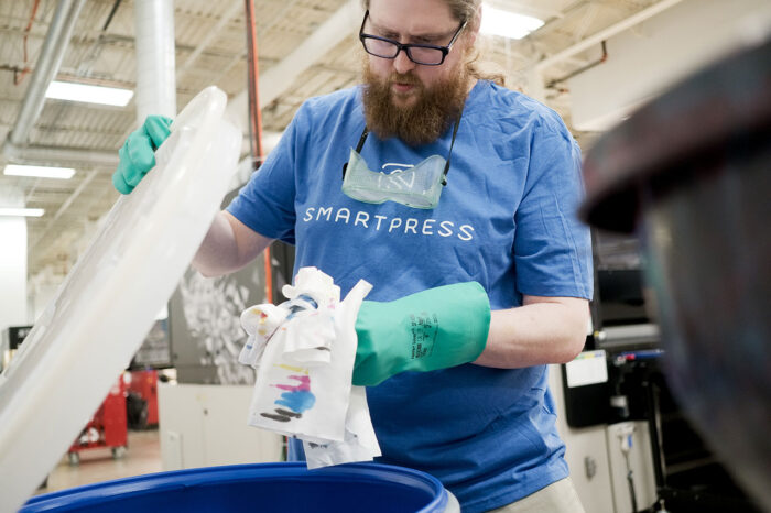 A man in a Smartpress T-shirt throwing ink-stained paper towels into a blue bin in a production area.