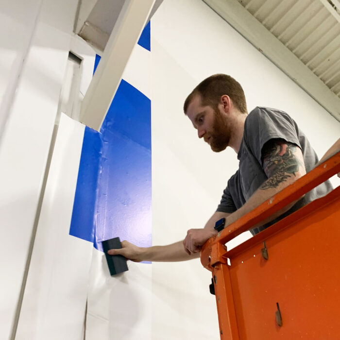 A man standing on an orange lift smoothing out a blue and white wall decal in a production facility.