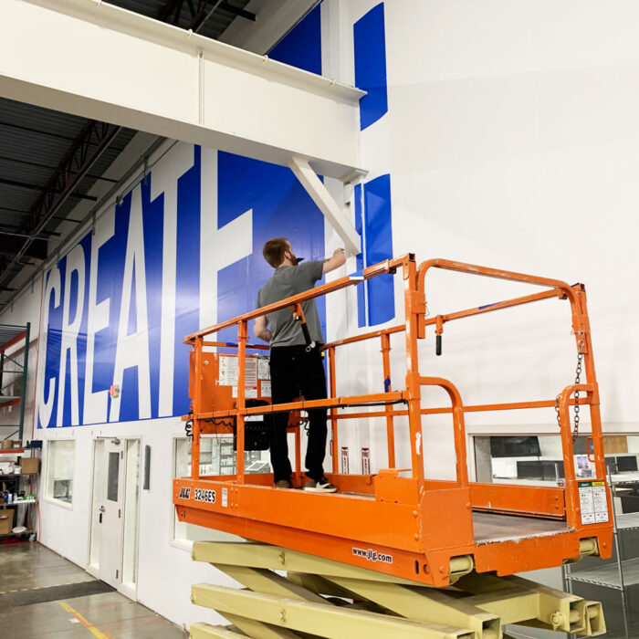 A man standing on an orange lift in a production facility installing a custom wall decal printed with Create in blue and white.