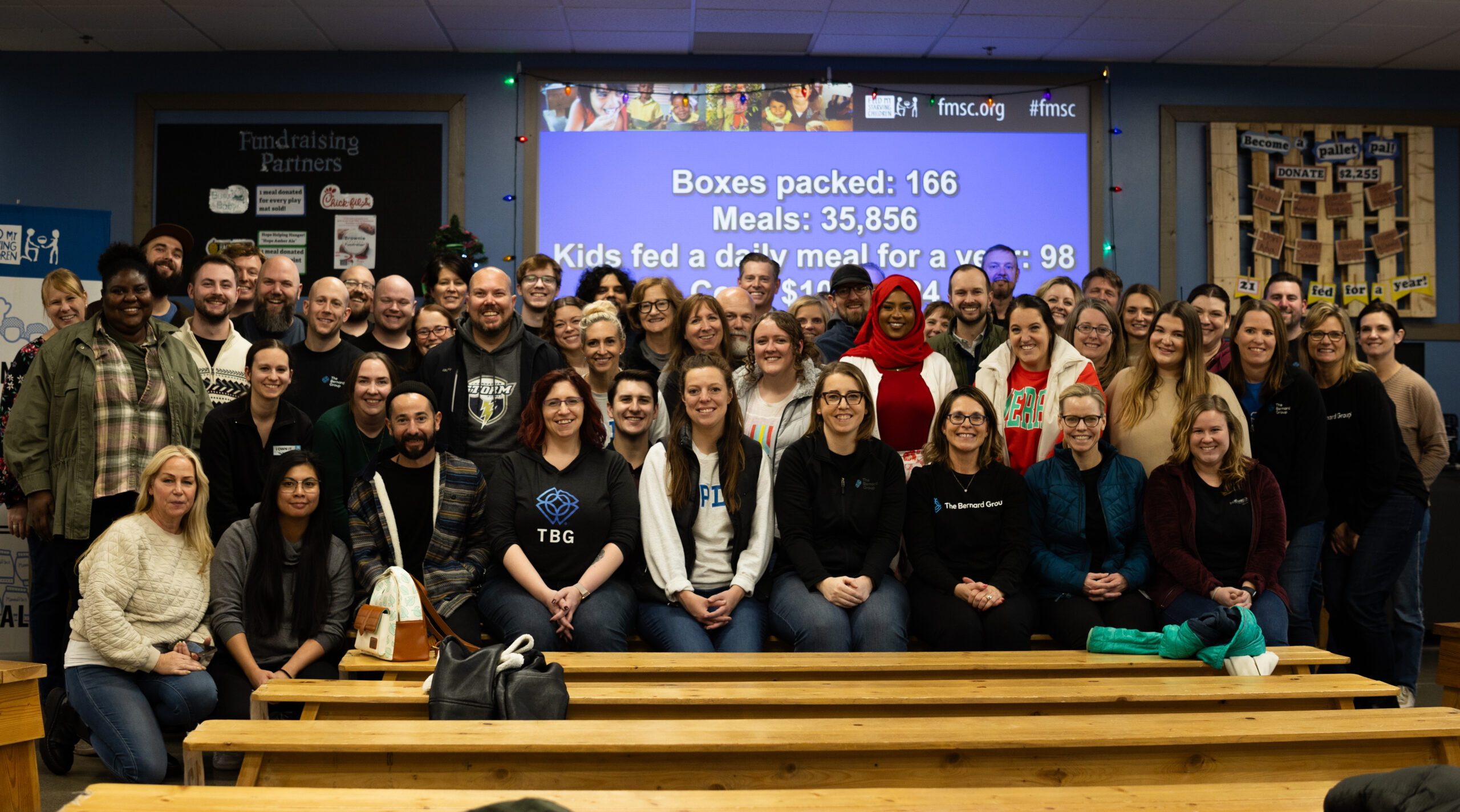 A group of people sitting and standing in rows in front of a screen that lists the number of boxes of food packed, meals and children fed at Feed My Starving Children.