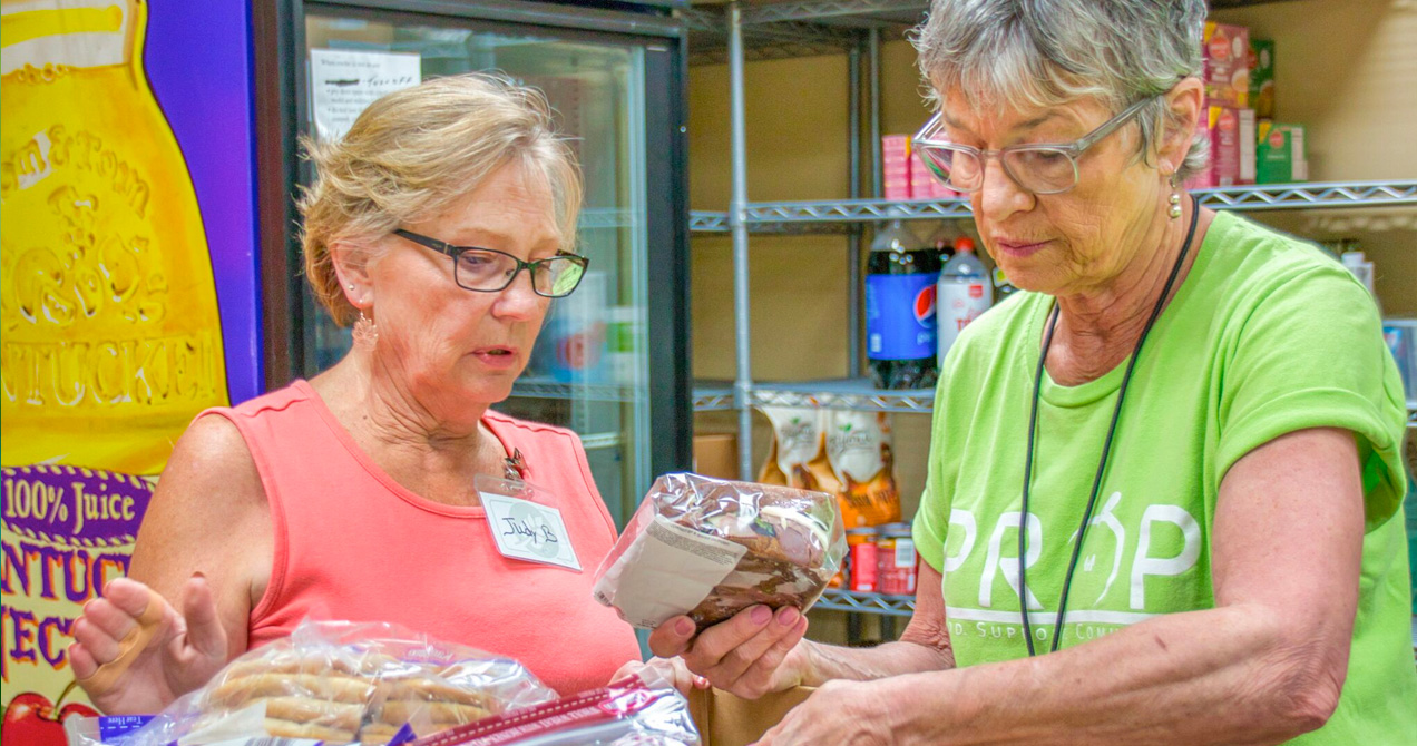 Two older women wearing glasses and sorting through packages of food for People Reaching Out to People.