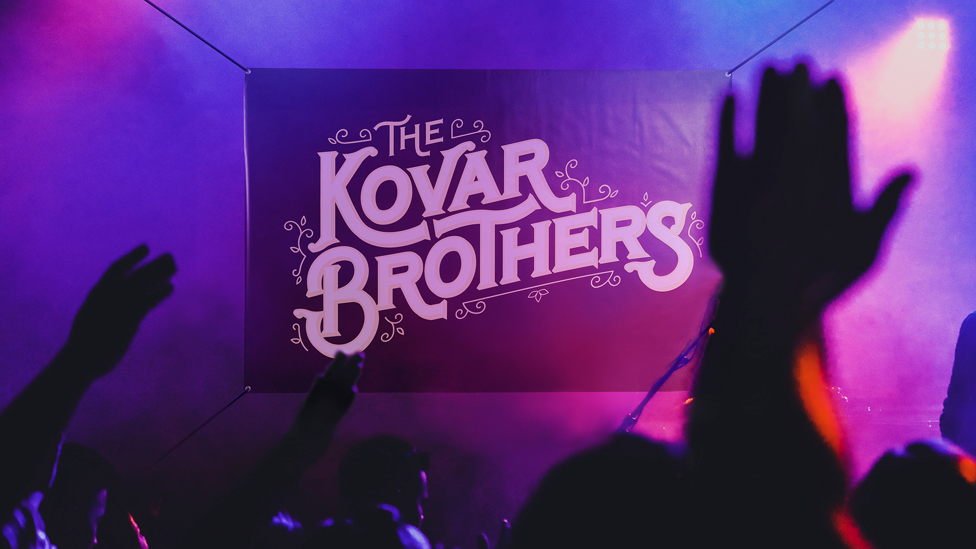 A custom banner printed with The Kovar Brothers in shades of pink and purple hanging in front of an audience.