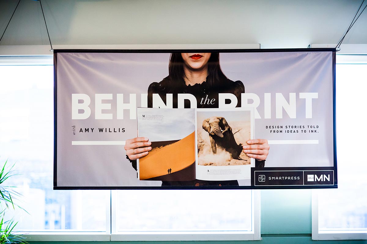 A hanging banner with Behind the Print in white text and a woman holding a booklet open.