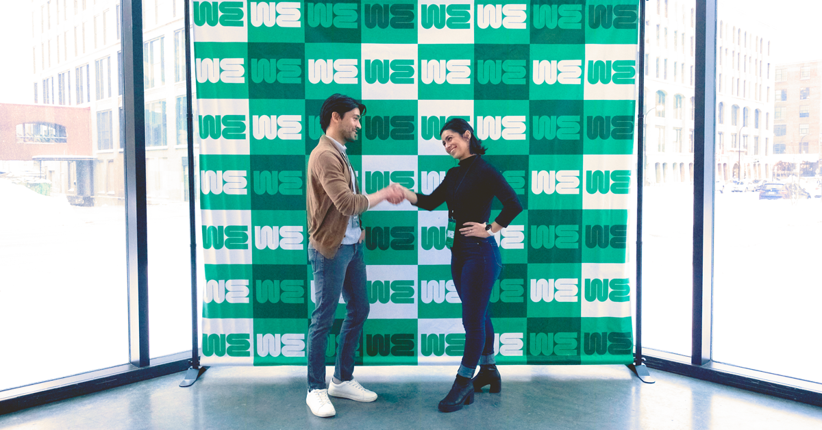 A man and woman shaking hands in front of a step and repeat banner printed with a green and white design.