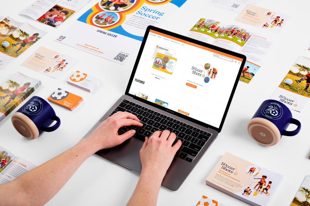 Handing typing on the laptop with the Soccer Shot web-to-print software displayed. Print materials are in the background displaying the vast marketing capacities.