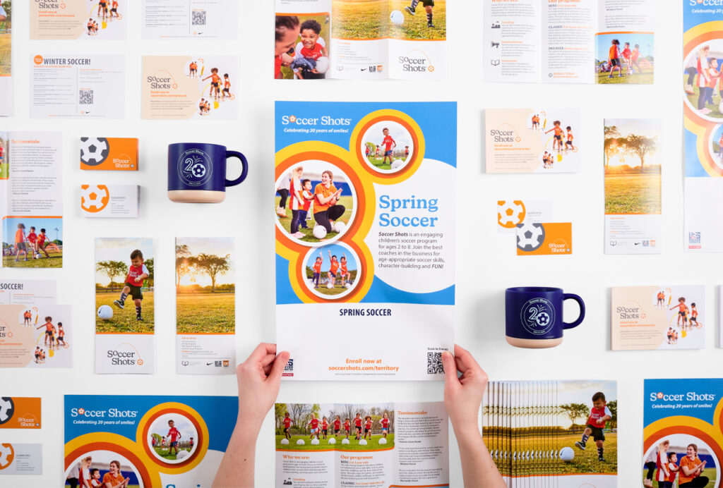 The Soccer Shots collective display of marketing collateral possible with web-to-print software. Two hands are holding a Soccer Shots flyer.