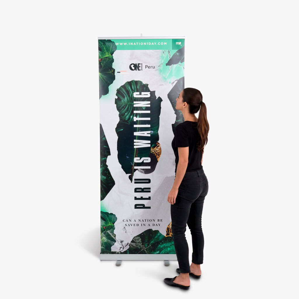 A hanging sign for 1 Nation 1 Day. Peru is waiting is spelling on vertically with plants in the background. There is a woman standing at side to show the height capabilities.