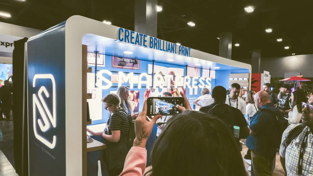 In the front is a woman taking a picture of the Smartpress booth. In the foreground, there are other people talking to Smartpress representatives at Adobe Max.