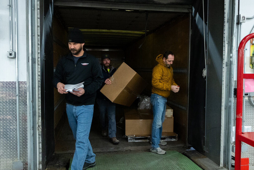 Three logistics professionals unloading large, heavy cardboard freight boxes from the back of an delivery truck container.
