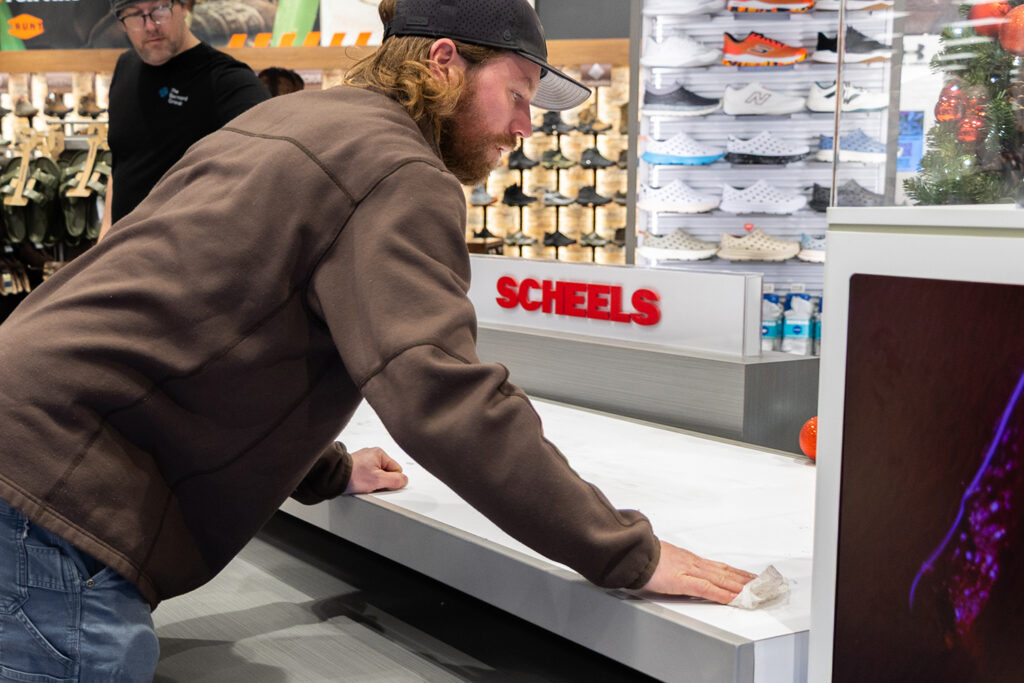 An installation technician carefully wiping down and prepping a clean white counter surface in front of an in-store Scheels retail department sign.