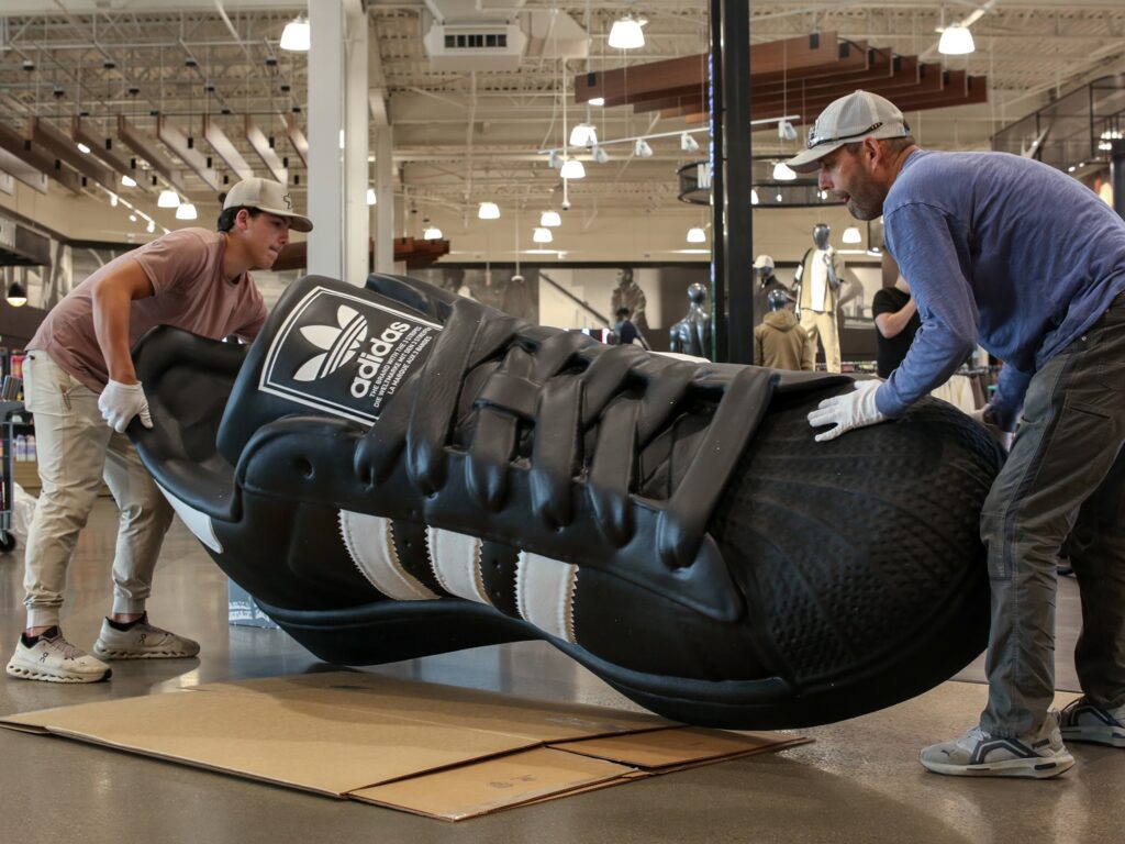 Two field installation professionals wearing protective gloves carefully position a massive, oversized Adidas sneaker display inside a sports apparel store.