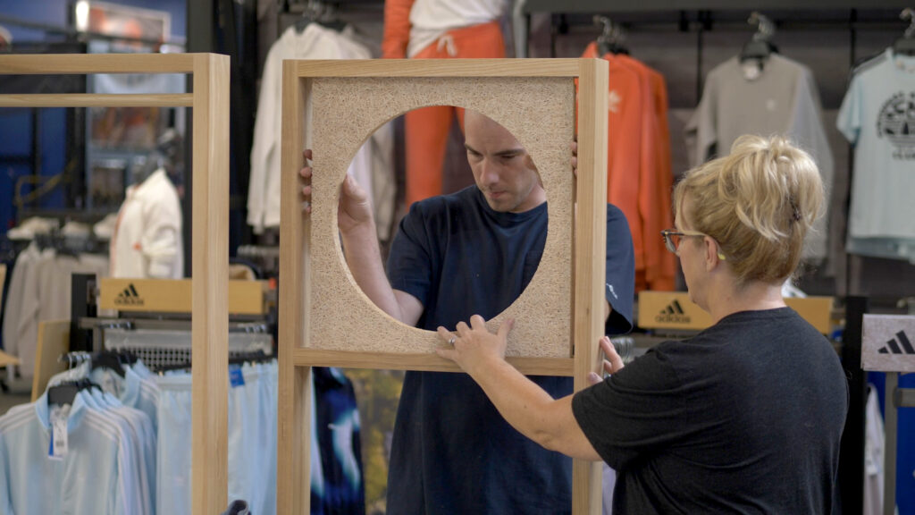 Two retail installation professionals aligning a wooden frame display with a circular cutout panel inside a clothing store.