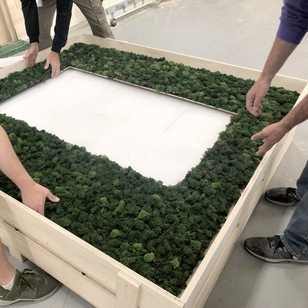 Close-up of workers meticulously placing organic green moss decor inside a custom wooden retail display frame.