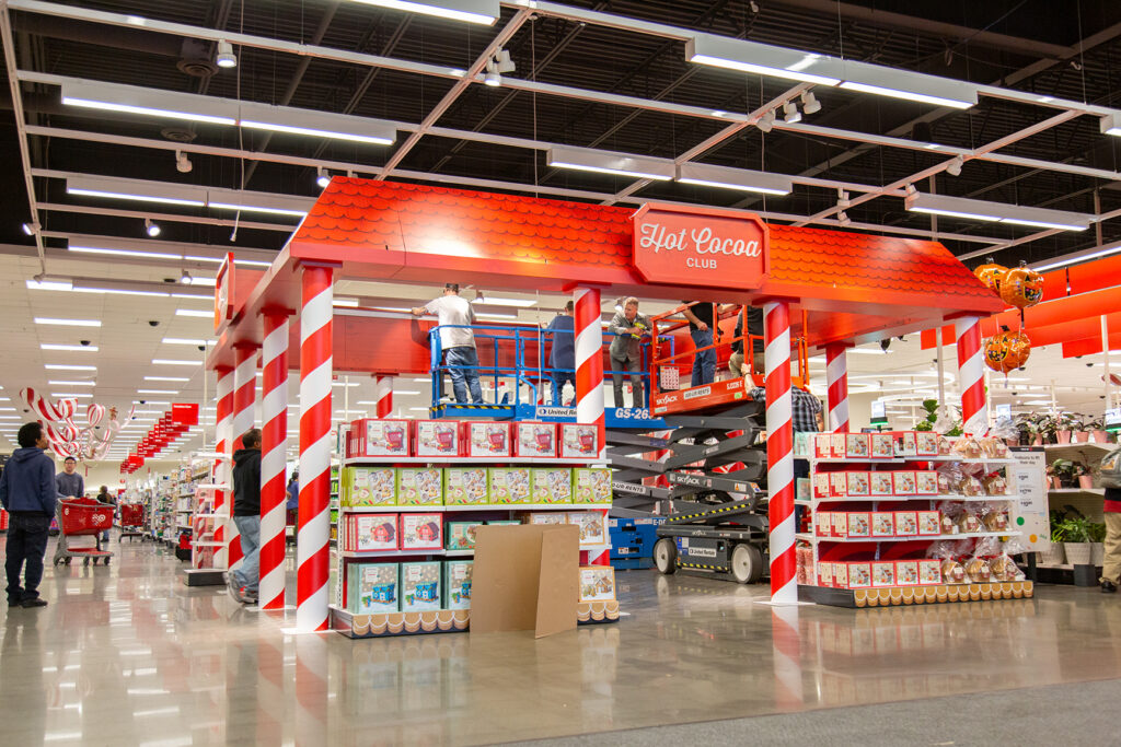 Wide view of a large candy-cane striped holiday "Hot Cocoa Club" pop-up structure being built in the center of Target using scissor lifts.