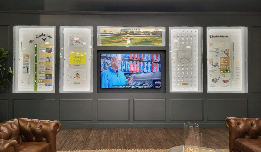 Experiential golf history lounge featuring leather sofas, illuminated glass display cases with Taylormade gear, and a central video display.