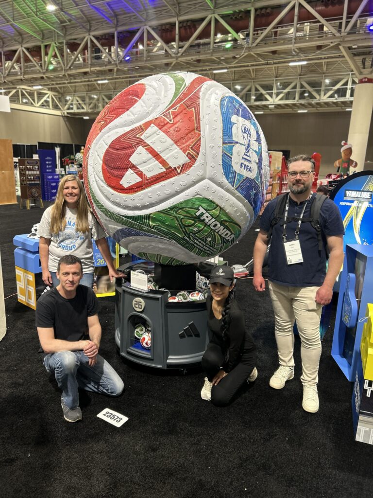 Four people posing around a giant 60-inch adidas World Cup 2026 soccer ball retail display featuring a gumball-machine design with real match balls stored inside the clear bins below.