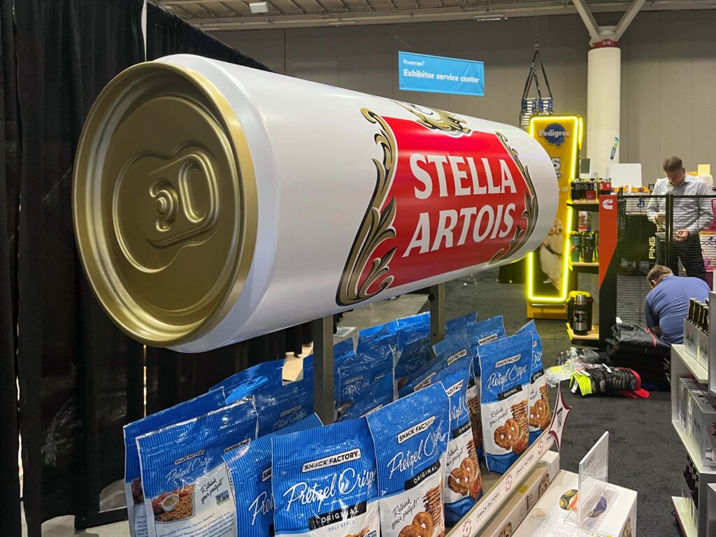 An oversized 3D molded replica of a Stella Artois slim can serving as a floor-standing retail fixture with cross-merchandising shelves.