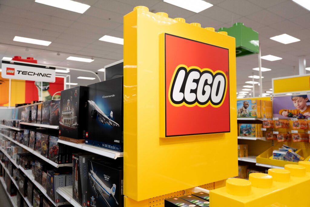Close-up of a vibrant yellow LEGO brick display fixture featuring the red LEGO logo on a retail aisle endcap, with Technic sets on nearby shelving.