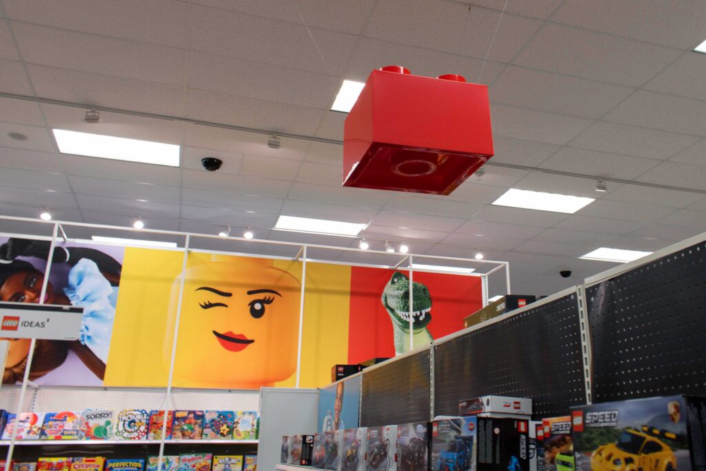 Overhead view of a large red LEGO brick fixture suspended from the ceiling above the retail aisle.
