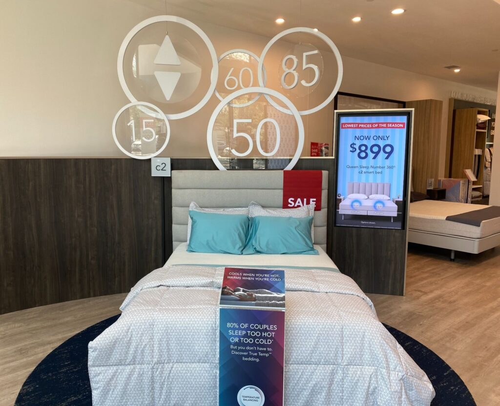 Sleep Number retail showroom display featuring a dressed smart bed with temperature messaging in a comfortable, semi-private setting.