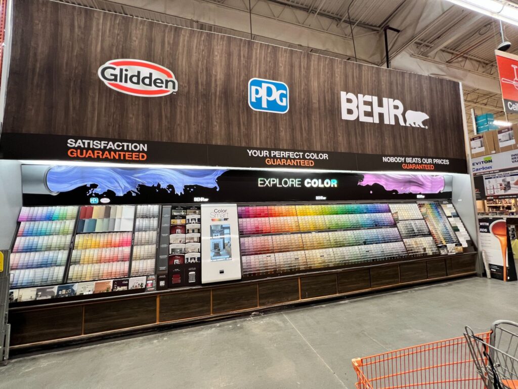 Large wood-grain wall signage featuring Glidden, PPG, and Behr logos above an organized retail paint department shelf display to guide customer navigation.