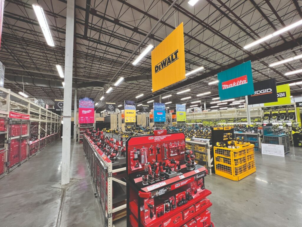 High-visibility yellow DEWALT and blue Makita brand banners hanging high above hardware store aisles to serve as visual spatial anchors for shoppers.