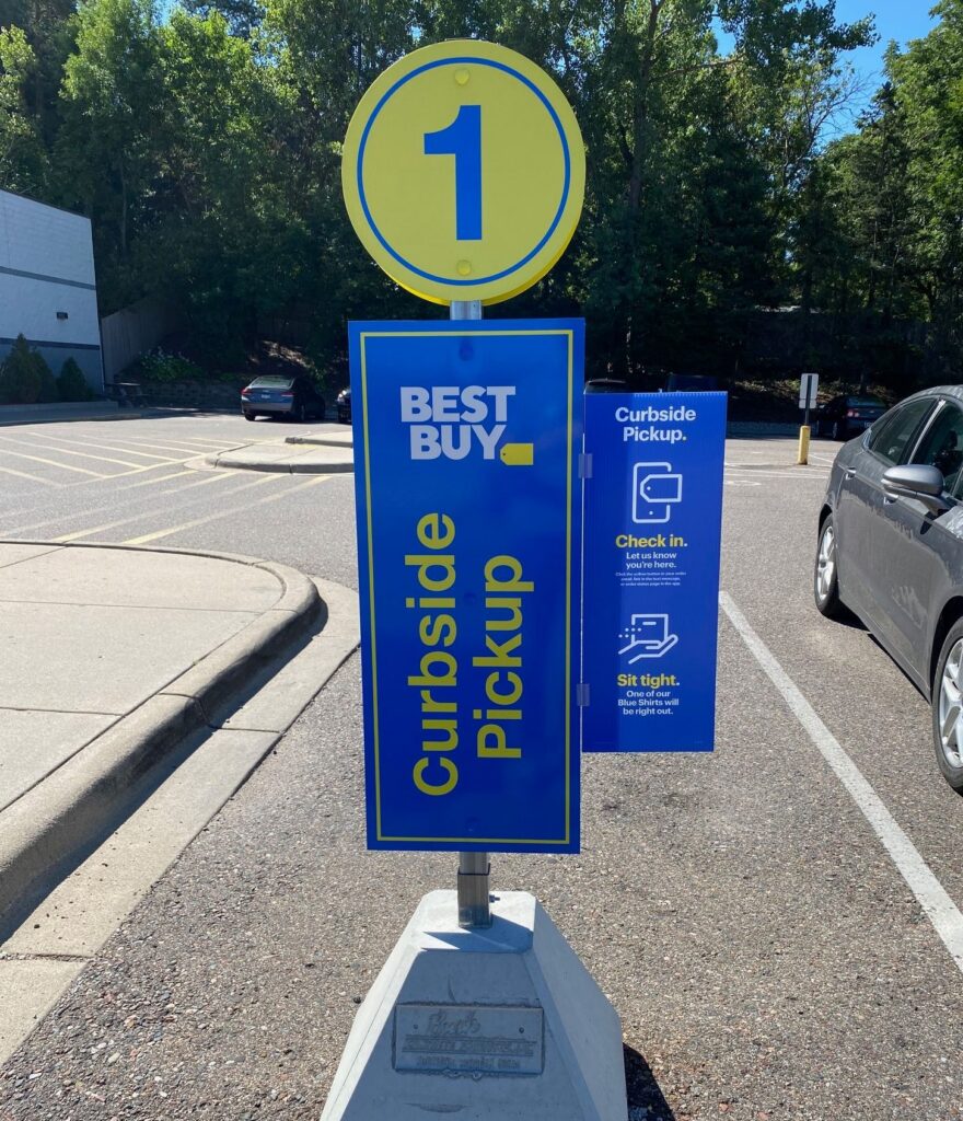 Weather-resistant blue and yellow Best Buy Curbside Pickup stanchion sign in a retail store parking lot.